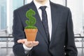 A person in formal suit holds a flowerpot with grass green dollar sign.