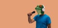 Person in frog mask with a microphone in hand and wireless headphones singing and enjoying karaoke on orange background