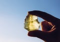 Person hand holding one yellow green lemon quartz crystal stone against sun and blue sky.