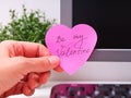 A person hand sticking a pink heart shaped paper note with words Be My Valentine on it on to a monitor at an office workplace Royalty Free Stock Photo