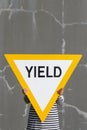 Person holds up big yield road sign. Bold yellow against cracked gray concrete wall.