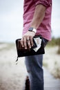 A person holding a black thick notebook, passport and brown wallet with money