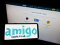 Person holding cellphone with business logo of British loan guarantor Amigo Holdings PLC on screen in front of webpage.