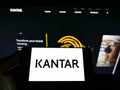 Person holding cellphone with business logo of British market research company Kantar Group on screen in front of webpage.