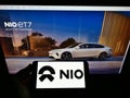Person holding cellphone with business logo of Chinese electric car manufacturer NIO Inc. on screen in front of webpage.