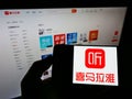 Person holding cellphone with business logo of Chinese online radio platform Ximalaya on screen in front of webpage.