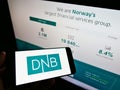 Person holding cellphone with business logo of Norwegian financial services company DNB ASA on screen in front of web page.