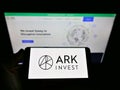 Person holding cellphone with company logo of US asset manager ARK Investment Management LLC on screen in front of webpage.