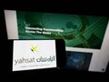 Person holding cellphone with logo of Al Yah Satellite Communications Company (Yahsat) on screen in front of webpage.