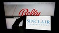 Person holding cellphone with logo of American company Sinclair Broadcast Group Inc. (SBG) on screen with webpage.