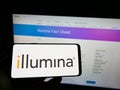 Person holding cellphone with logo of American genetics company Illumina Inc. on screen in front of business webpage.
