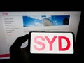 Person holding cellphone with logo of Australian company Sydney Airport Holdings Pty Ltd on screen in front of webpage.