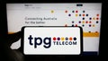 Person holding cellphone with logo of Australian company TPG Telecom Limited on screen in front of business webpage.