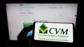 Person holding cellphone with logo of Brazilian Comissao de Valores Mobiliarios (CVM) on screen in front of webpage. Royalty Free Stock Photo