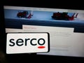 Person holding cellphone with logo of British service company Serco Group plc on screen in front of business webpage.