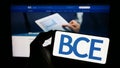 Person holding cellphone with logo of Canadian telecommunications company BCE Inc. on screen in front of webpage.