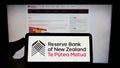 Person holding cellphone with logo of central bank Reserve Bank of New Zealand (RBNZ) on screen in front of webpage.