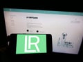 Person holding cellphone with logo of cosmetics company LR Health and Beauty Systems GmbH on screen in front of webpage.