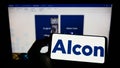 Person holding cellphone with logo of eye care company Alcon AG on screen in front of business webpage.
