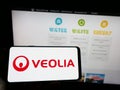 Person holding cellphone with logo of French company Veolia Environnement SA on screen in front of business webpage.