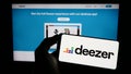 Person holding cellphone with logo of French music streaming company Deezer S.A. on screen in front of business webpage.