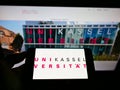 Person holding cellphone with logo of German university UniversitÃÂ¤t Kassel on screen in front of webpage.