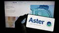 Person holding cellphone with logo of health company Aster DM Healthcare Limited on screen in front of business webpage.