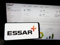 Person holding cellphone with logo of Indian conglomerate Essar Group on screen in front of business webpage.