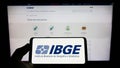 Person holding cellphone with logo of Instituto Brasileiro de Geografia e Estatistica (IBGE) on screen with webpage.