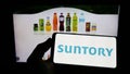 Person holding cellphone with logo of Japanese beverage company Suntory Holdings K.K. on screen in front of webpage.