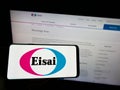 Person holding cellphone with logo of Japanese pharmaceutical company Eisai Co Ltd on screen in front of business webpage.