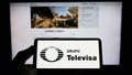 Person holding cellphone with logo of Mexican media company Grupo Televisa SAB on screen in front of business logo.