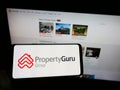 Person holding cellphone with logo of online property company PropertyGuru Pte. Ltd. on screen in front of webpage.