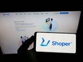 Person holding cellphone with logo of Polish software company Shoper S.A. on screen in front of business webpage.