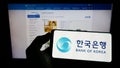 Person holding cellphone with logo of South Korean central bank Bank of Korea (BOK) on screen in front of webpage.