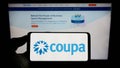 Person holding cellphone with logo of spend management company Coupa Software Inc. on screen in front of business webpage.