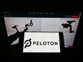 Person holding cellphone with logo of US company Peloton Interactive Inc. on screen in front of business webpage.