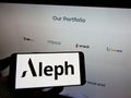 Person holding cellphone with logo of US digital media company Aleph Holding on screen in front of business webpage.