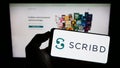 Person holding cellphone with logo of US publishing platform company Scribd Inc. on screen in front of business webpage.