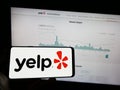 Person holding cellphone with logo of US review platform company Yelp Inc. on screen in front of business webpage.