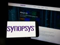 Person holding cellphone with logo of US technology company Synopsys Inc. on screen in front of business webpage.