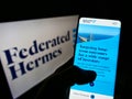 Person holding cellphone with web page of US investment manager Federated Hermes Inc. on screen in front of logo.