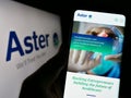 Person holding cellphone with webpage of health company Aster DM Healthcare Limited on screen in front of logo.