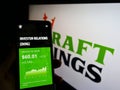 Person holding cellphone with webpage of US sports betting company DraftKings Inc. on screen in front of logo.