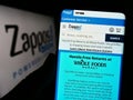 Person holding cellphone with website of US online shop company Zappos.com LLC on screen in front of logo.