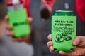 Person holding a green beer can in a green koozie with another person holding the same in the background at a festival