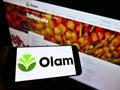 Person holding mobile phone with logo of agribusiness company Olam International Limited on screen in front of webpeg.