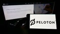 Person holding mobile phone with logo of American company Peloton Interactive Inc. on screen in front of business web page.