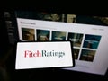 Person holding mobile phone with logo of American credit rating agency Fitch Ratings Inc. on screen in front of webpage.