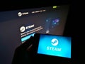 Person holding mobile phone with logo of American distribution platform Steam operated by Valve on screen in front of website.